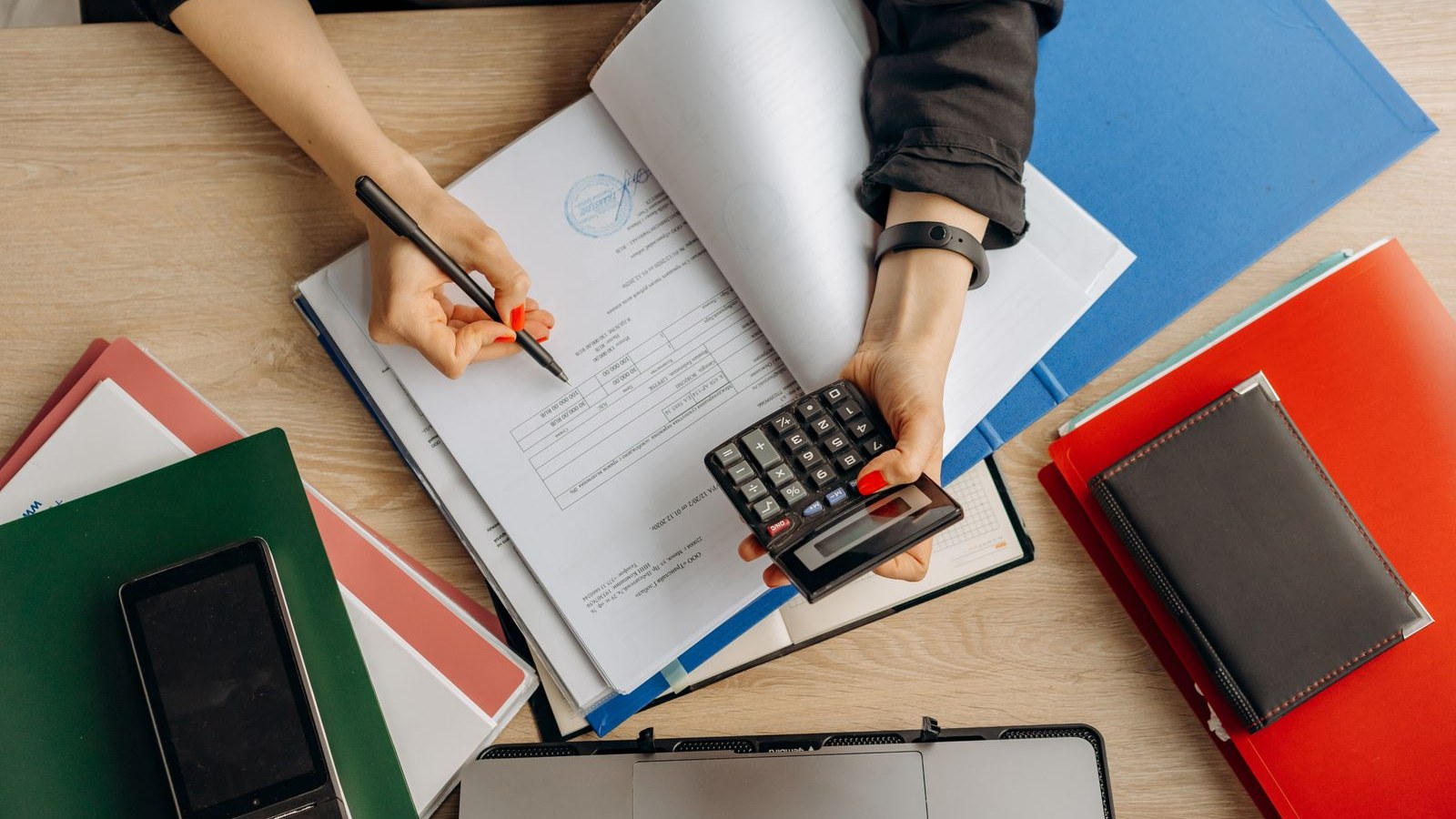 Photo d’illustration : une personne vérifie des documents avec une calculatrice à son bureau.